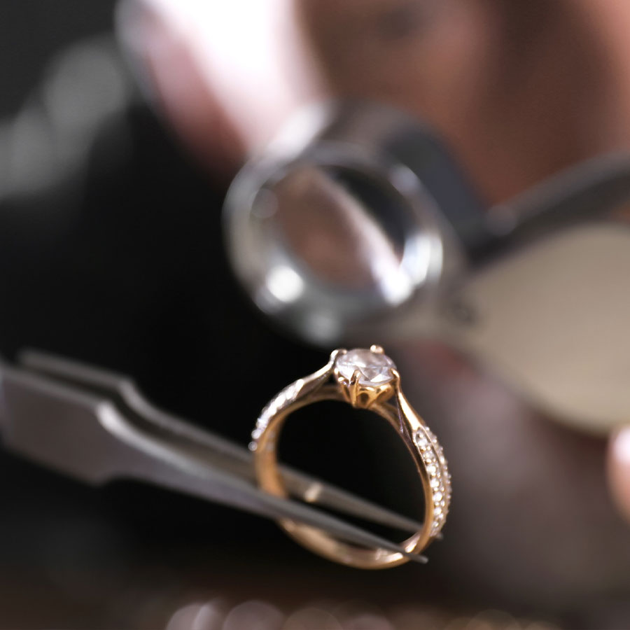 A closeup of a jeweler holding long tweezers to appraise a gold diamond engagement ring.